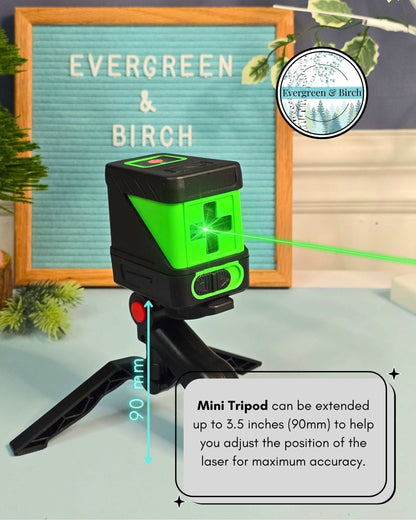 Green self-leveling laser level mounted on adjustable mini tripod projecting horizontal beam for precise gallery wall frame alignment.