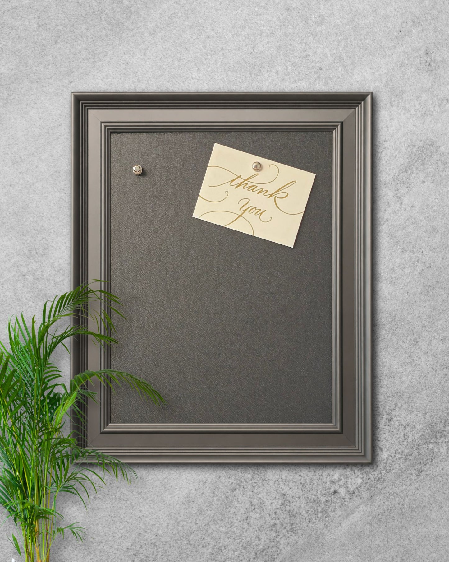 Modern black magnetic board with solid wood frame mounted on a textured gray wall, featuring a pinned “thank you” note and decorative plant styling.