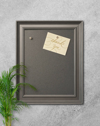 Modern black magnetic board with solid wood frame mounted on a textured gray wall, featuring a pinned “thank you” note and decorative plant styling.