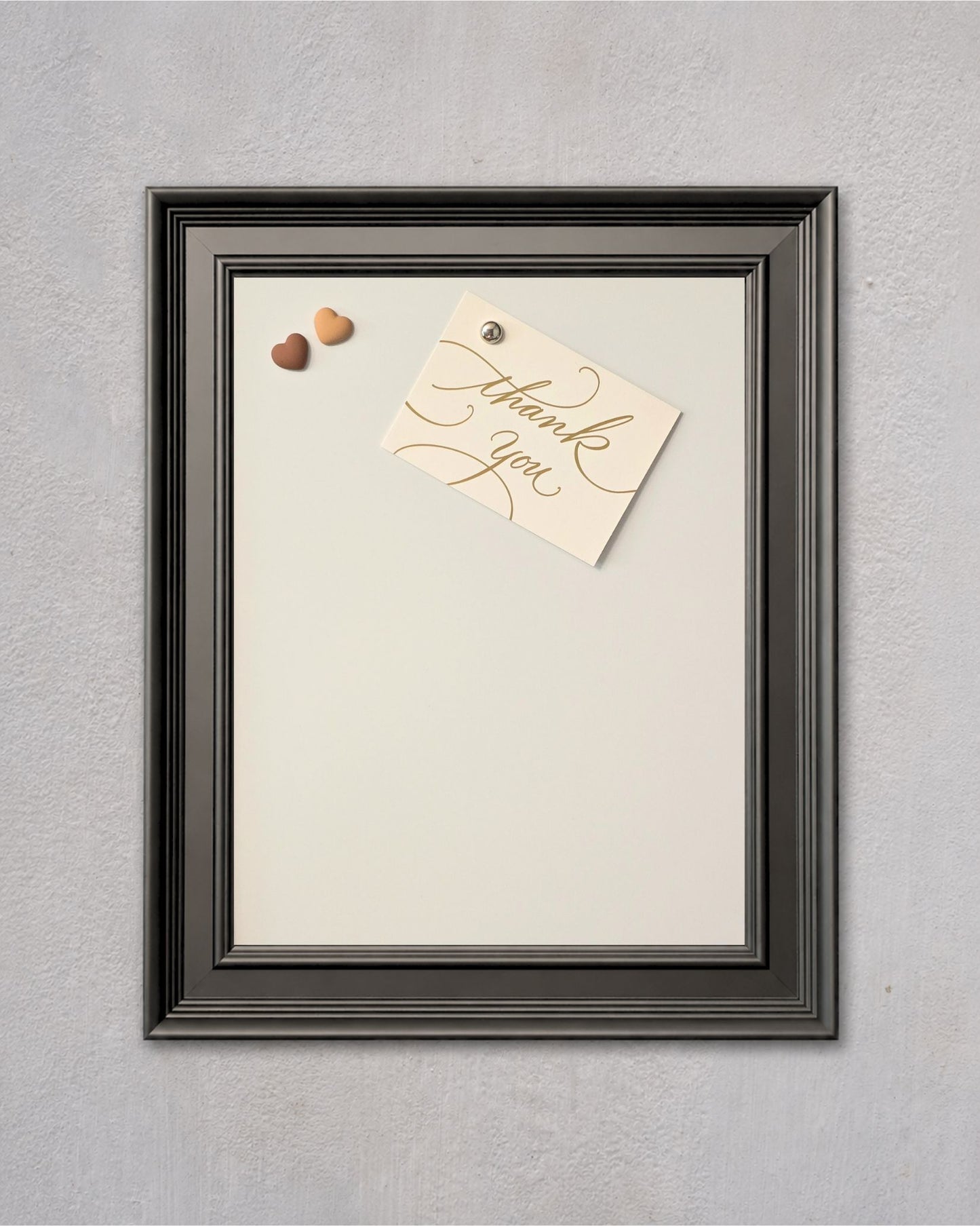 Modern black magnetic board with white powder-coated steel surface, featuring a pinned thank you note and decorative heart magnets on a textured gray wall.