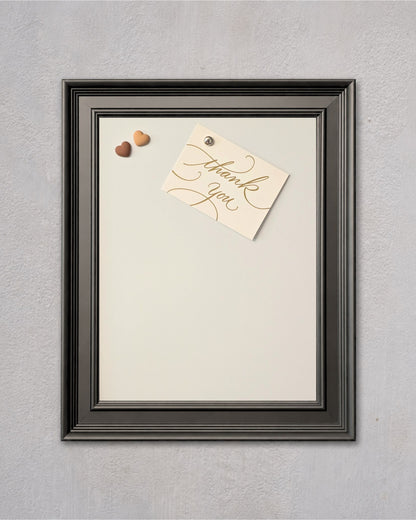 Modern black magnetic board with white powder-coated steel surface, featuring a pinned thank you note and decorative heart magnets on a textured gray wall.